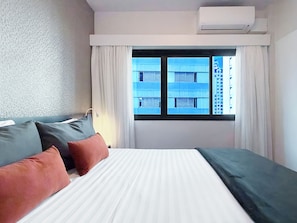 A hotel room with a bed, pillows, an air conditioning unit, a window with curtains, and a view of buildings outside.