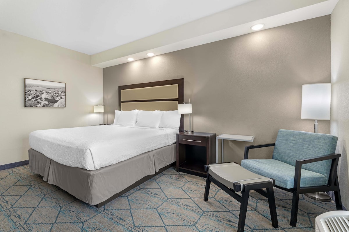 A hotel room with a large bed, a chair, a small table, and a nightstand with a lamp.