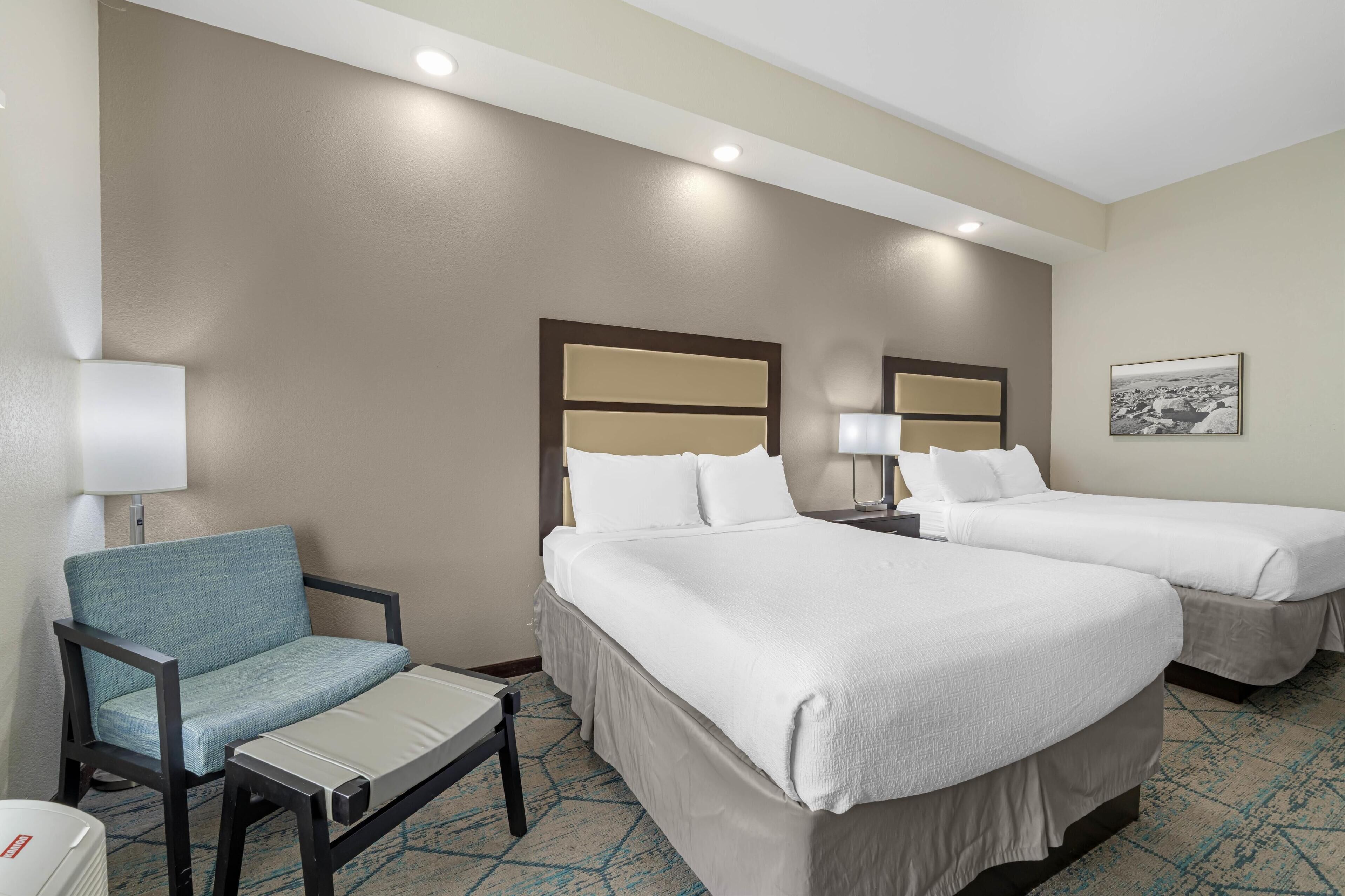 A hotel room with two beds, a chair, a small table, and a lamp.