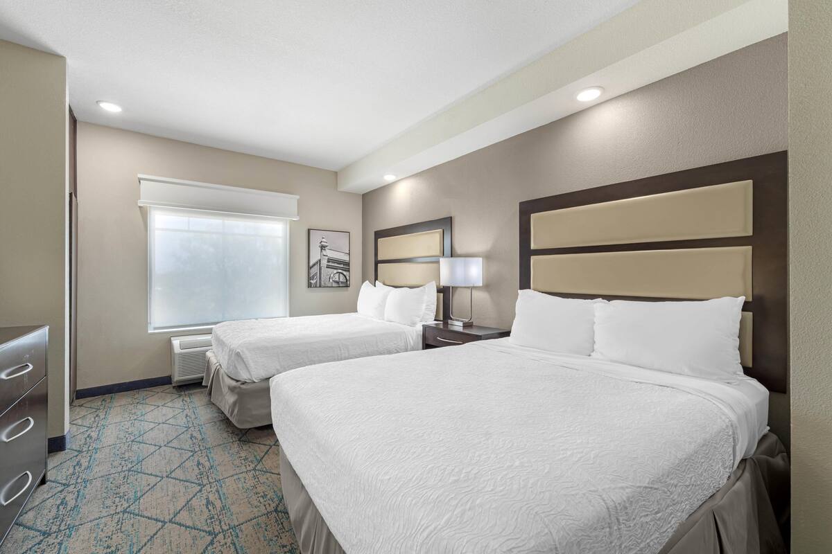 A hotel room with two beds, a dresser, a window with blinds, a framed picture on the wall, and a patterned carpet.