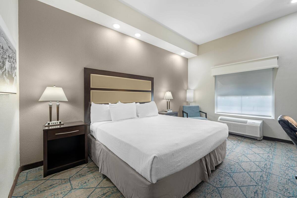A hotel room with a large bed, two bedside tables with lamps, a window with blinds, and a wall-mounted air conditioner.