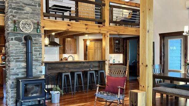 A spacious living area with a stone fireplace, wooden beams, a kitchen with a bar, and a high ceiling. - Custom Finger Lakes home near Ithaca with hot tub  (Newfield)