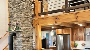 A rustic kitchen with a stone wall, wooden beams, a black stove, and a bar area with stools. - Custom Finger Lakes home near Ithaca with hot tub  (Newfield)