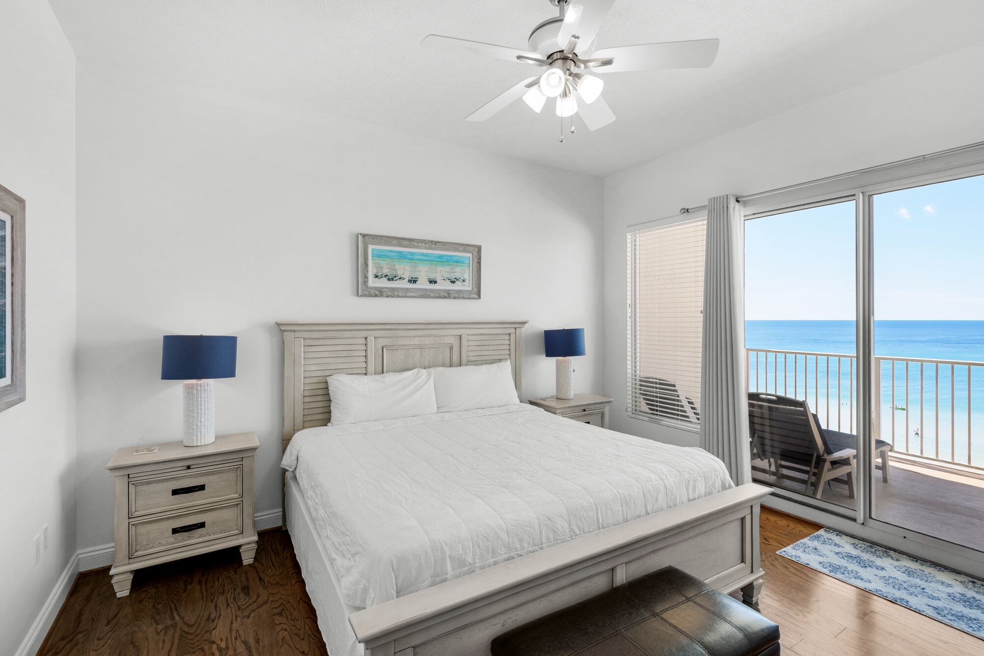 A bedroom with a bed, two bedside tables with lamps, a ceiling fan, a large window with a view of the ocean, and a balcony with a chair.