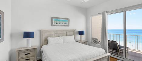 A bedroom with a bed, two bedside tables with lamps, a ceiling fan, a large window with a view of the ocean, and a balcony with a chair.