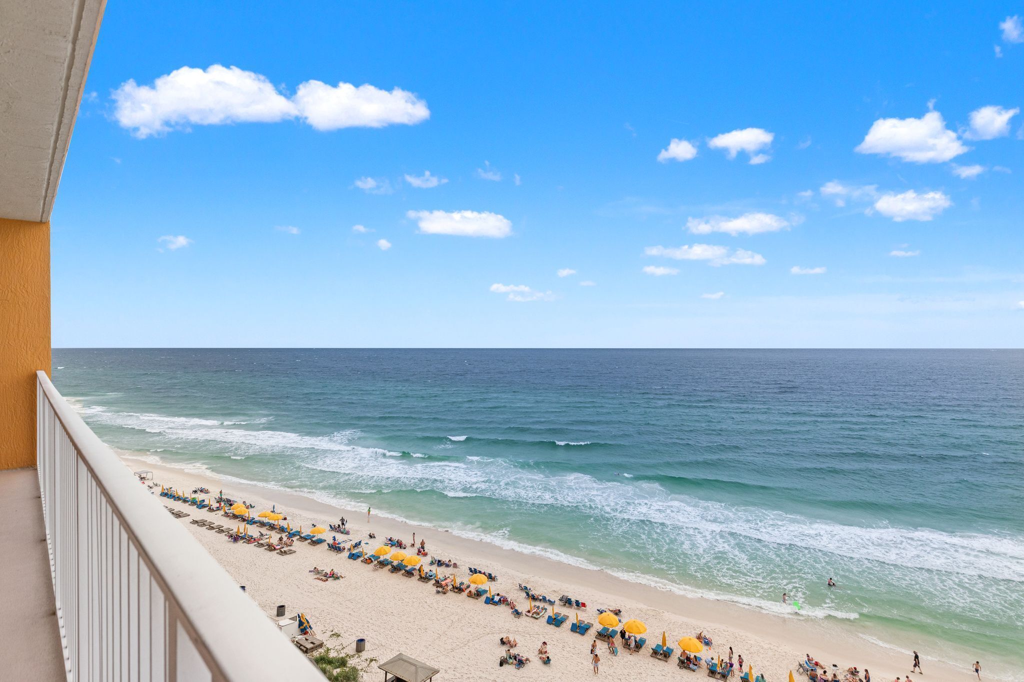 A beach view from a balcony with a clear sky, ocean waves, and beach umbrellas.