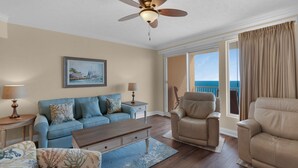 A living room with a sofa, armchairs, a coffee table, and a ceiling fan. There is a painting on the wall and a view of the ocean through the sliding glass doors. - Treasure Island 1611 (Panama City Beach)