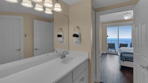 A bathroom with a large mirror, a sink, and towel hooks. There is a door leading to a balcony with a view of the ocean. - Treasure Island 1611 (Panama City Beach)