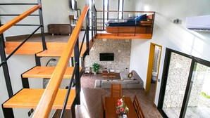 Un apartament modern d'estil loft amb una escala de fusta, un llit de paleria, zona de dinar i un saló amb sofà i televisió.