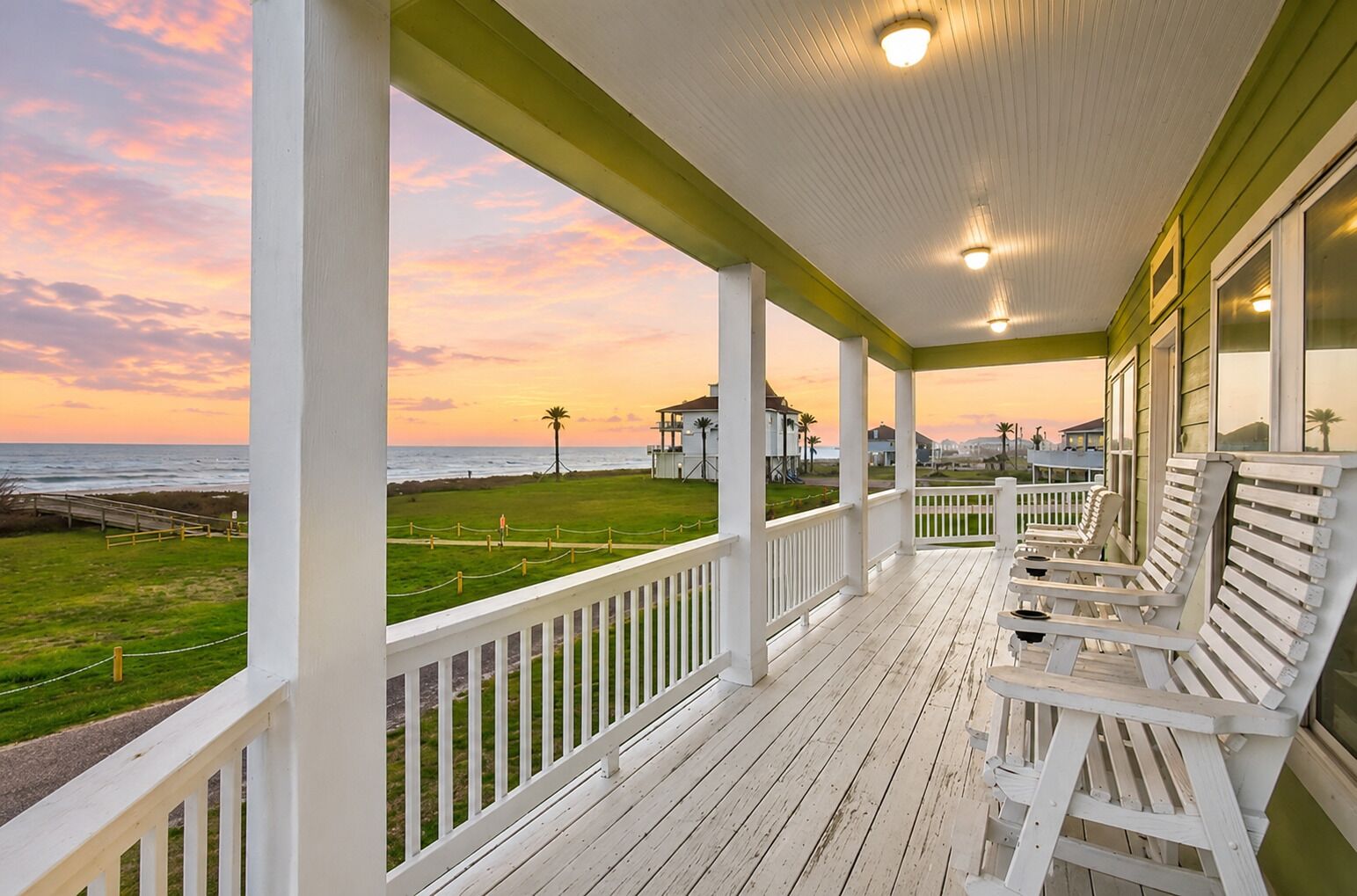 A covered porch with white railings and benches, overlooking a grassy area with a view of the ocean and buildings in the distance.