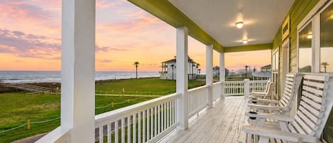 A covered porch with white railings and benches, overlooking a grassy area with a view of the ocean and buildings in the distance.