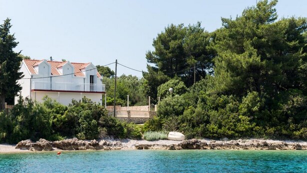 A coastal building with a red-tiled roof, surrounded by dense green trees and a clear blue sea. - Holiday Home Maćus-One Bedroom Apartment with Balcony (Kolocep)