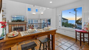 A dining area with a wooden table set for two, a fireplace, and a large window with a view of the ocean. - Spanish Palms | 3BR Home Steps to Windansea Beach (San Diego)