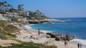 A coastal beach with people sunbathing and swimming, houses on a hillside, and rocky shoreline. - Spanish Palms | 3BR Home Steps to Windansea Beach (San Diego)