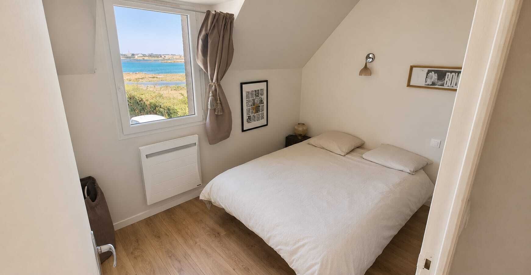 A bedroom with a bed, a window with a view of the sea, a radiator, and a framed picture on the wall.