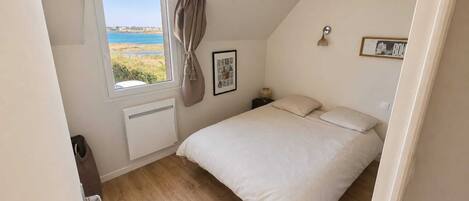 A bedroom with a bed, a window with a view of the sea, a radiator, and a framed picture on the wall.