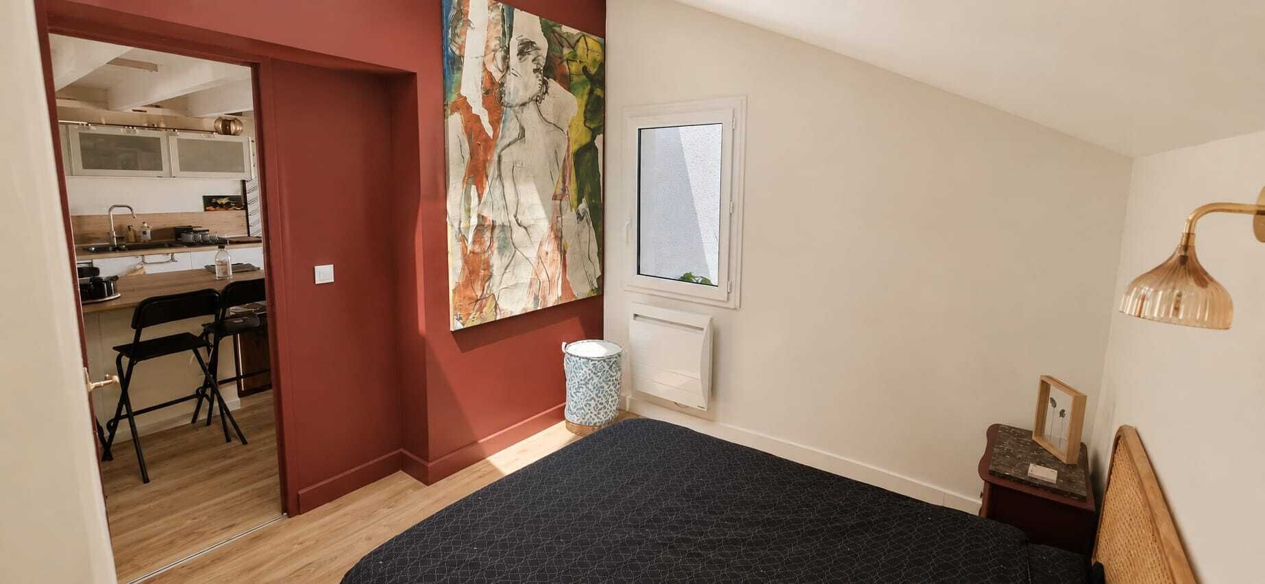 A bedroom with a bed, a nightstand, a lamp, a window, and a view of a kitchen area.