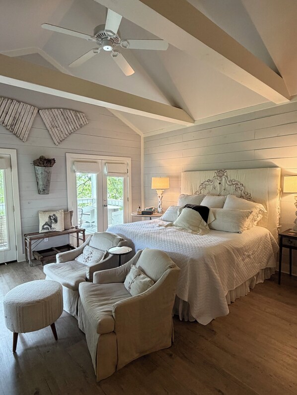A bedroom with a large bed, two chairs, a ceiling fan, and a window with a view.