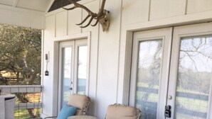 A covered outdoor seating area with wicker chairs and a table, a deer skull mounted on the wall, and a glass door leading outside. - Cozy and comfortable one bedroom with kitchen and with pasture view. (Burton)