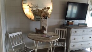 A room with a round mirror, a small table with chairs, a vase with dried flowers, a television, and a dresser with drawers. - Cozy and comfortable one bedroom with kitchen and with pasture view. (Burton)