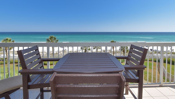 Un balcon avec une table et deux chaises, donnant sur une plage aux eaux d’un bleu limpide et sur une côte lointaine.