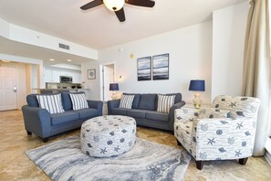 Ein Wohnzimmer mit Sofa, Hocker und Sesseln, einem Deckenventilator und einer Küche im Hintergrund.