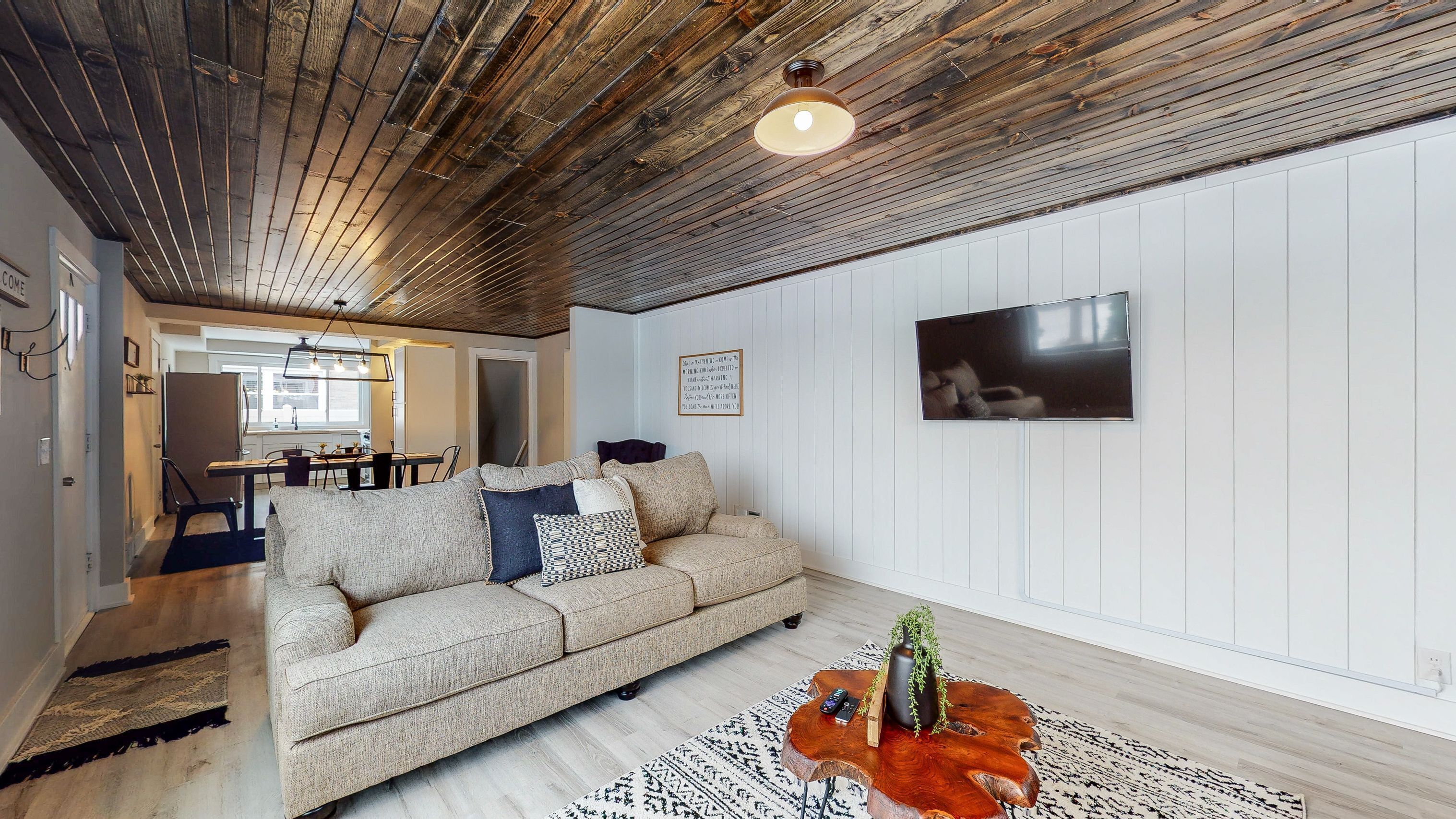 Una sala de estar con techo de madera, un sofá color beige, un televisor montado en la pared y un comedor al fondo.
