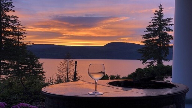 A glass of white wine on a round table with a view of a lake and mountains during sunset.