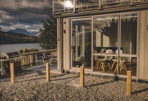 Un bâtiment moderne doté d’une porte et de fenêtres en verre, d’un sol en gravier et offrant une vue sur des montagnes et un lac.