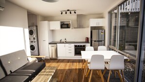 A modern kitchen with white cabinetry, a microwave, oven, and a refrigerator. There is a dining area with a table and chairs, and a sofa with a wooden floor.