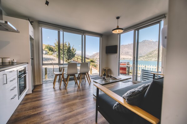 Une cuisine moderne avec un plancher en bois, un coin repas avec des chaises, un canapé et de grandes fenêtres offrant une vue sur un paysage montagneux.