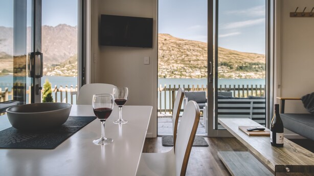 A dining area with a table, two wine glasses, a bowl, and a bottle of wine. The room has a large window with a view of mountains and a body of water.