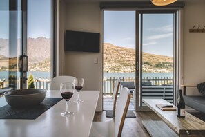 Un coin repas avec une table, deux verres à vin, un bol et une bouteille de vin. La pièce est dotée d’une grande fenêtre offrant une vue sur des montagnes et un plan d’eau.