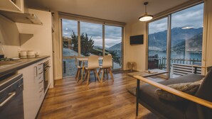 A modern kitchen with wooden flooring, a dining area with a table and chairs, and a sofa. Large windows offer a view of mountains and a lake.