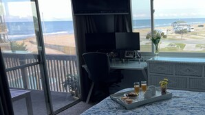 A room with a large window overlooking a beach, a desk with a chair, a bed with a floral bedspread, and a tray with breakfast items.