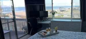 Une chambre dotée d’une grande fenêtre donnant sur une plage, d’un bureau avec un ordinateur et d’un plateau de petit-déjeuner sur le lit.