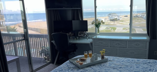 A room with a large window overlooking a beach, a desk with a chair, a bed with a floral bedspread, and a tray with breakfast items.