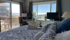 A hotel room with a bed, a desk with a computer, a TV, and a balcony with a view of the beach.