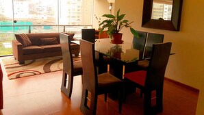 A modern living room with a glass dining table, dark chairs, a sofa, and a large window offering a view of buildings and greenery. - Nice Apartment Malecon Balta Miraflores (Lima)