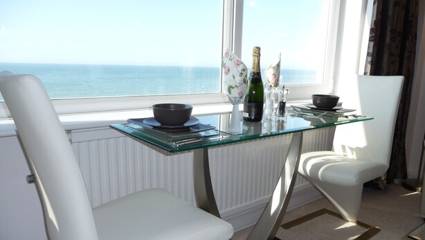 A modern dining area with a glass table, white chairs, a view of the sea, and a bottle of champagne. - With spectacular sea views the flat is literally across the road from the Beach. (Hove)
