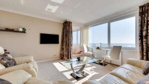 A modern living room with a sofa, a coffee table, and a TV. There is a window with curtains and a view of the sky. - With spectacular sea views the flat is literally across the road from the Beach. (Hove)