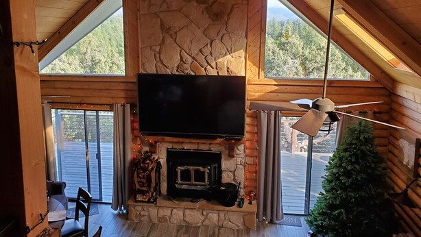 A cozy cabin interior with a stone fireplace, a television mounted above it, and large windows offering a view of the outdoors. - Stunning Views from 3 Level Home, Majestic Sunrise and Mountain Scapes (Pine)