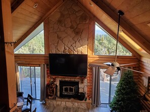 A cozy cabin interior with a stone fireplace, a television mounted above it, and large windows offering a view of the outdoors. - Stunning Views from 3 Level Home, Majestic Sunrise and Mountain Scapes (Pine)