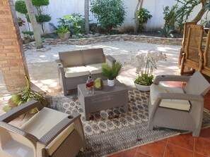 Relax in this serene outdoor seating area with a sofa, armchairs, and a small table, surrounded by lush greenery and a beautifully paved space.