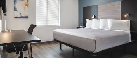 A modern hotel room with a large bed, a desk, and a window with blinds.