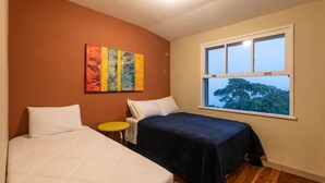 A bedroom with two beds, a small table, a window with a view of trees, and a colorful abstract painting on the wall. - Duplex apartment in a guesthouse in the Bocaina mountains with a view of the lake. (Bananal)