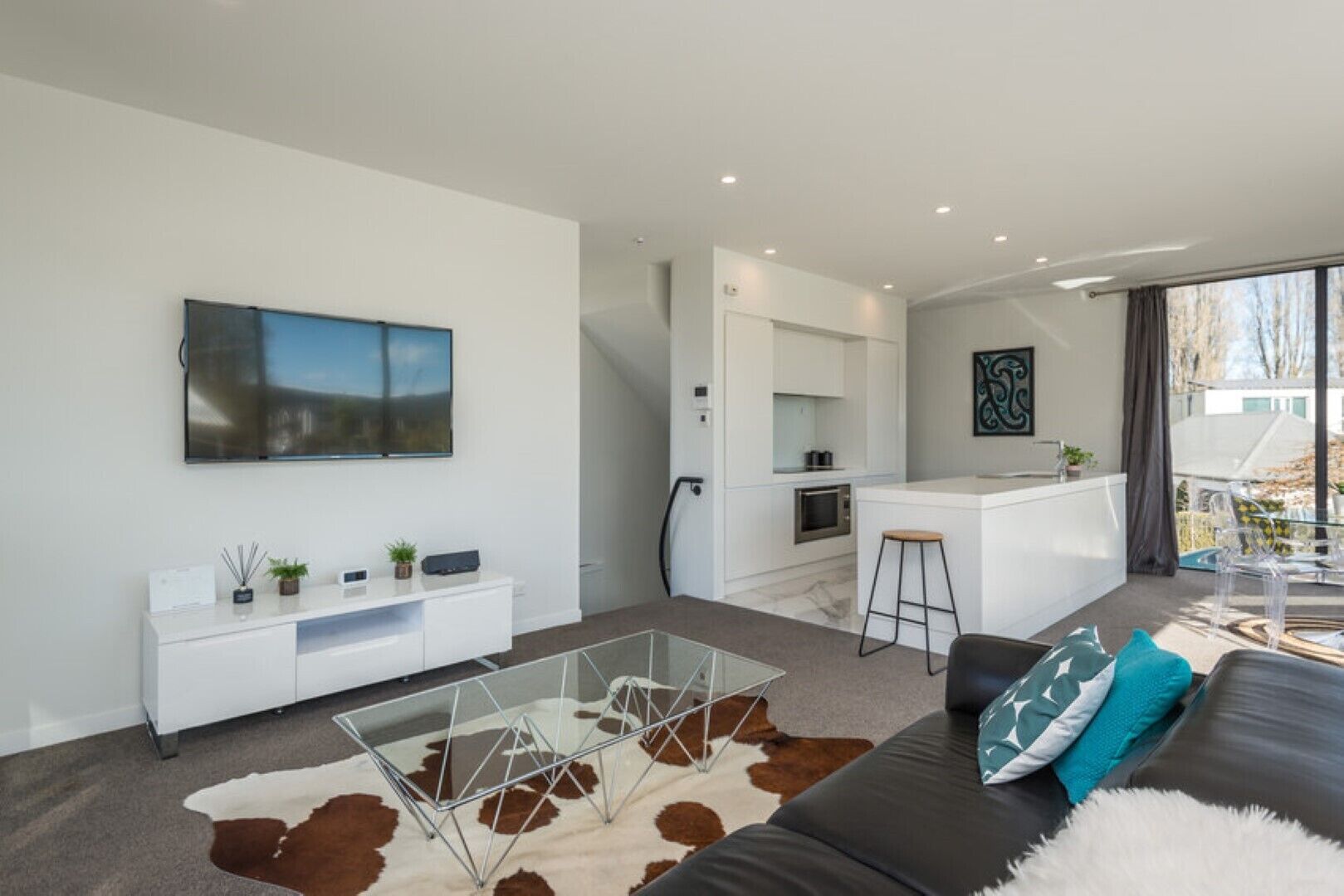 A modern living room with a glass coffee table, a cowhide rug, and a black sofa with blue and white pillows. A kitchen with white cabinets and a bar area is visible in the background.