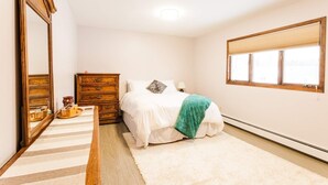 Quarto com um espelho grande, cômoda de madeira, cama com roupa de cama branca e manta verde, e janela com persianas.