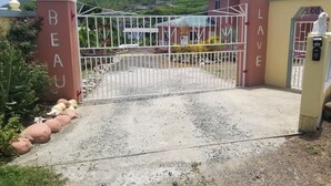 A gated entrance with "BEAU" and "LAVE" signs, a gravel driveway, and a building with a green roof in the background.