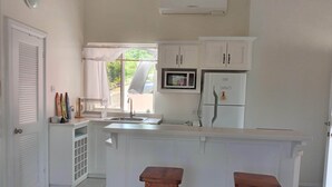 A kitchen with a white countertop, a sink, a microwave, and a refrigerator. There are two wooden stools and a window with a view of greenery outside.